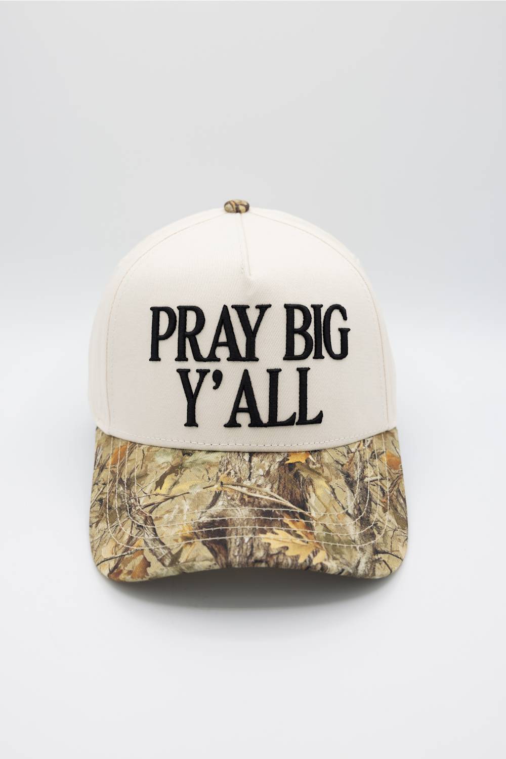 A baseball cap with an off-white crown and a Realtree camouflage patterned visor, featuring the text 'PRAY BIG Y'ALL' embroidered in black on the front.