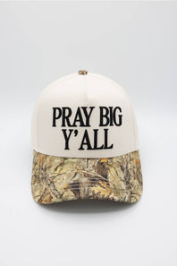 A baseball cap with an off-white crown and a Realtree camouflage patterned visor, featuring the text 'PRAY BIG Y'ALL' embroidered in black on the front.
