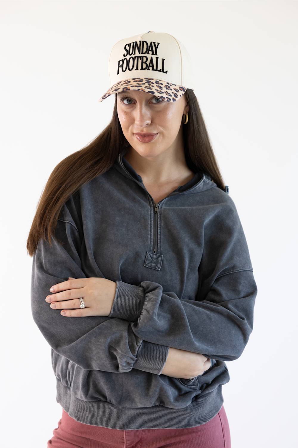 A woman wearing a washed charcoal gray quarter-zip sweatshirt, a cream and leopard-print 'SUNDAY FOOTBALL' cap, and burgundy pants, standing against a plain white background.