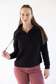 A woman modeling a black long-sleeve quarter-zip pullover sweatshirt with a mock neck, paired with dusty rose pants, against a plain white studio background.