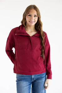 A woman modeling a burgundy half-zip pullover hoodie with a mock neck and front kangaroo pocket, paired with blue jeans, against a plain white studio background.