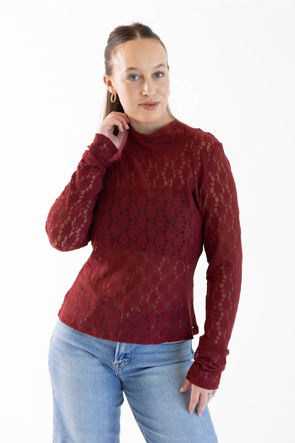A woman modeling a long-sleeve mock neck top in a deep burgundy lace fabric, paired with light blue jeans, against a plain white background.