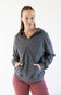 A woman modeling a dark gray washed half-zip hoodie with a drawstring hood and front kangaroo pockets, paired with dusty rose pants, against a plain white studio background.