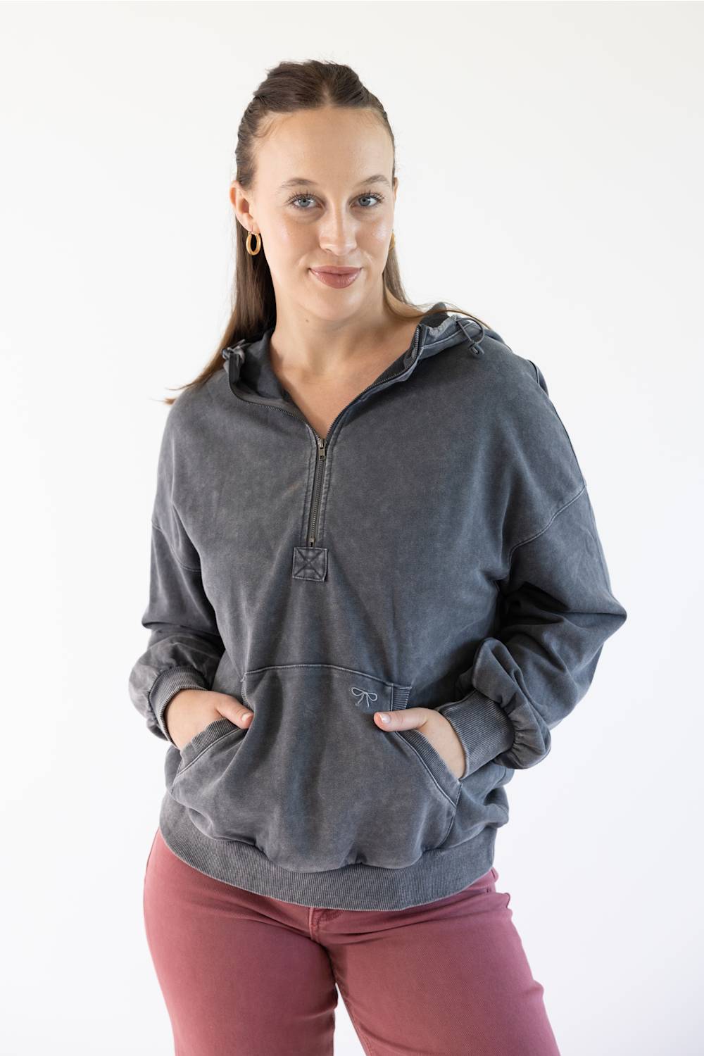 A woman modeling a dark gray washed half-zip hoodie with a drawstring hood and front kangaroo pockets, paired with dusty rose pants, against a plain white studio background.