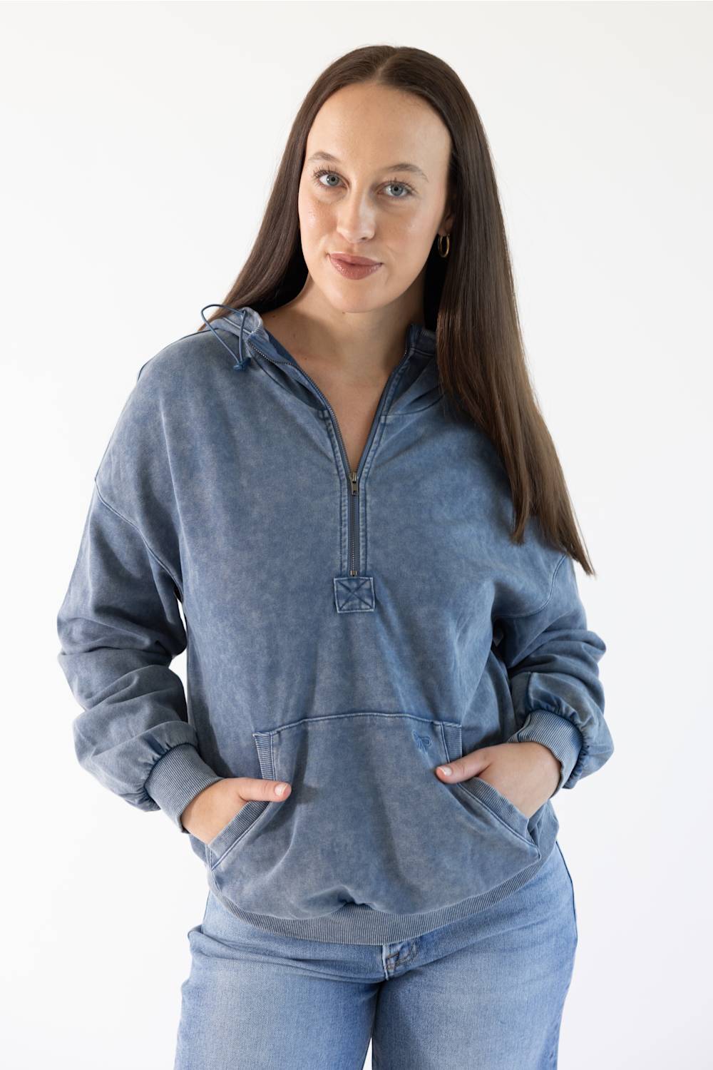 A woman modeling a washed blue half-zip hoodie with a drawstring hood and front kangaroo pockets, paired with blue jeans, against a plain white studio background.