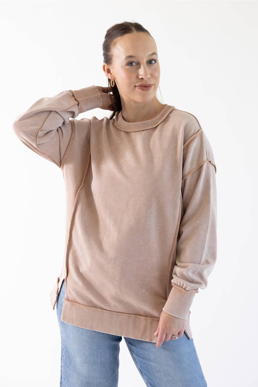 A woman modeling a light beige crewneck sweatshirt with long sleeves and side slits, paired with blue jeans, against a plain white background.