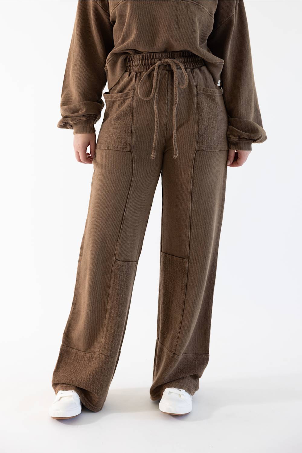 A person wearing loose brown drawstring sweatpants and a matching brown sweatshirt, standing against a plain white studio background with white sneakers visible.
