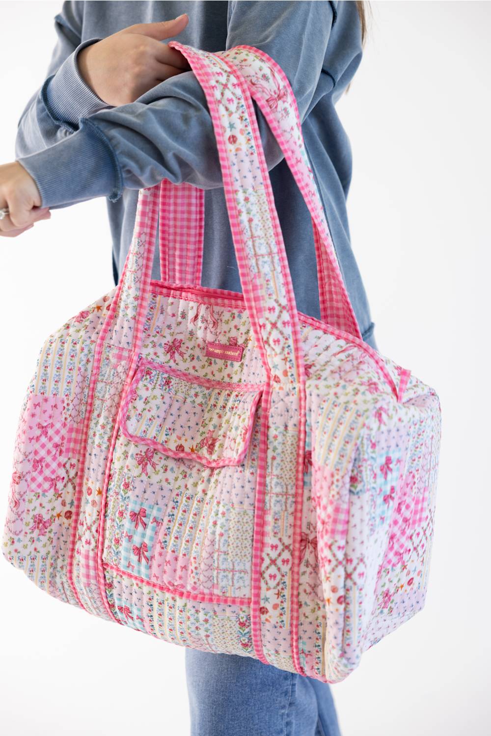 A person wearing a blue sweatshirt and jeans carries a large patchwork fabric duffel bag with pink gingham trim and floral prints on a white background.