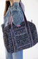 A person wearing a gray sweatshirt and blue jeans carries a large floral print duffel bag with purple trim and a patterned shoulder strap against a plain white background.