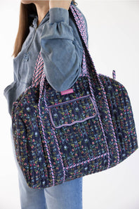 A person wearing a gray sweatshirt and blue jeans carries a large floral print duffel bag with purple trim and a patterned shoulder strap against a plain white background.