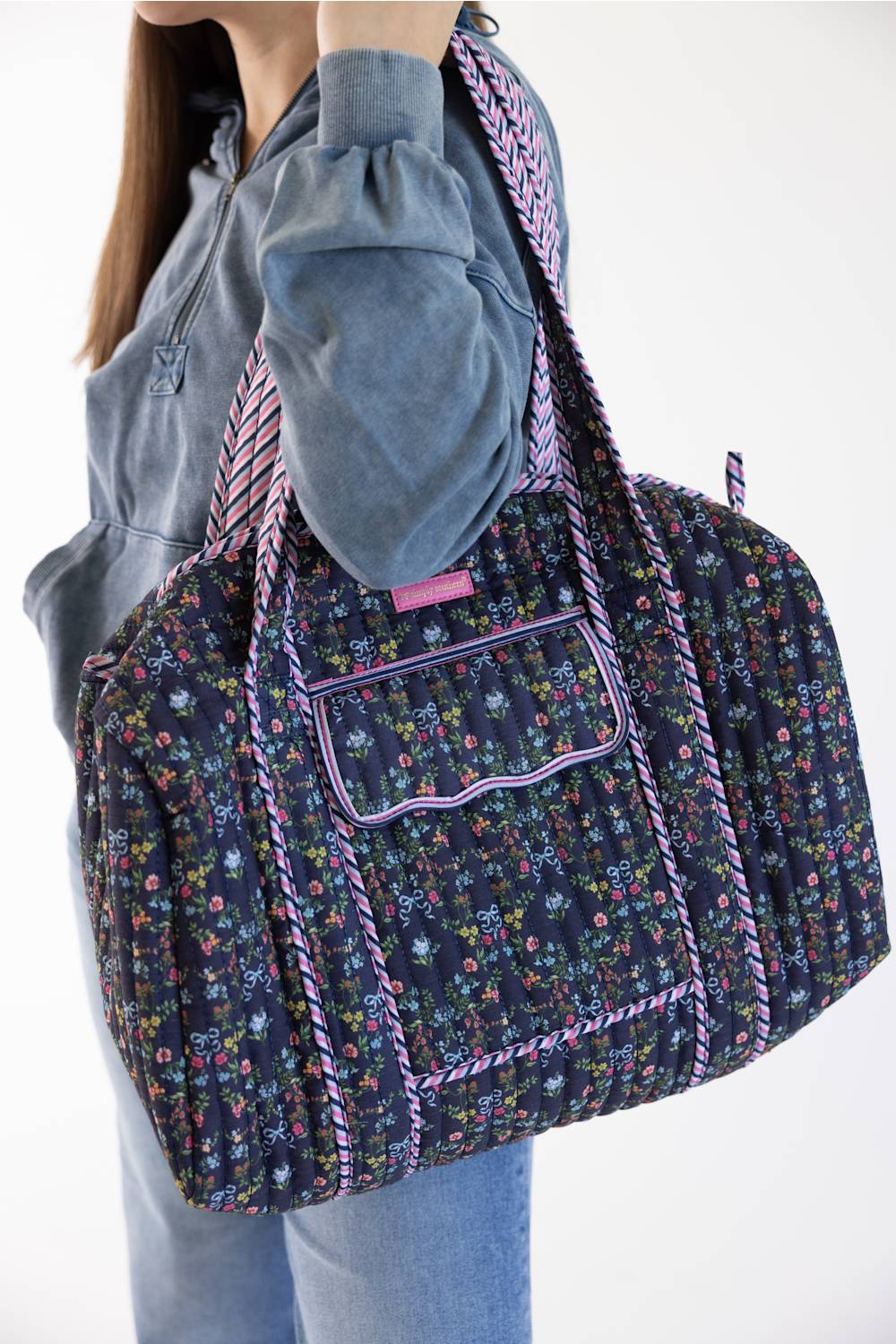 A person wearing a gray sweatshirt and blue jeans carries a large floral print duffel bag with purple trim and a patterned shoulder strap against a plain white background.