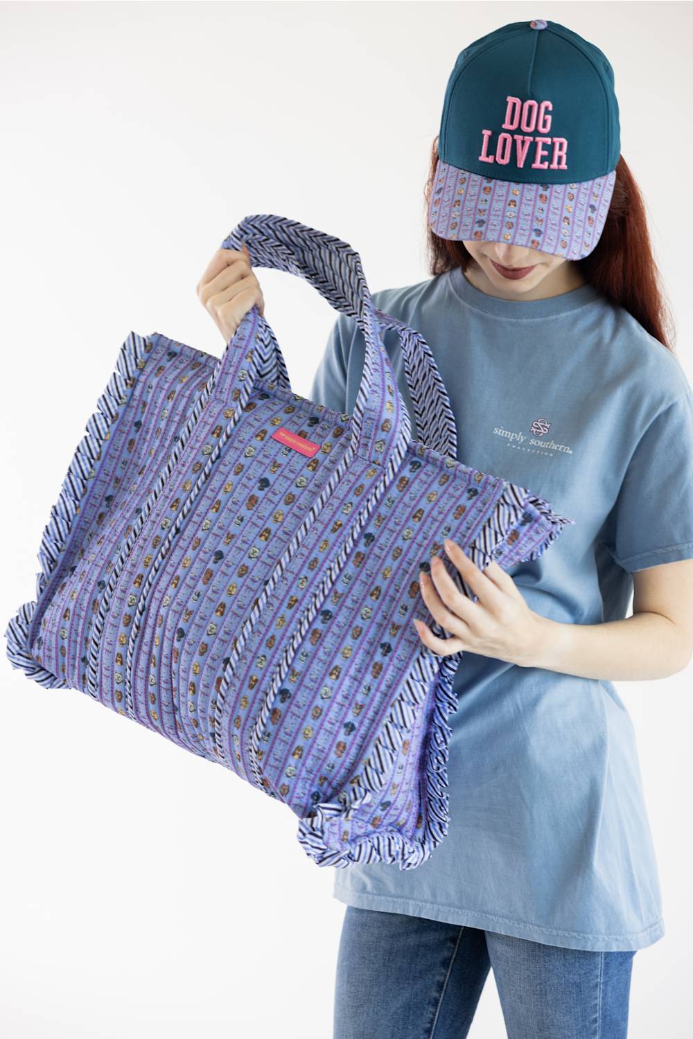 A person wearing a blue t-shirt and a teal cap with 'DOG LOVER' in pink text holds a large woven purple tote bag with fringe details against a plain white background.