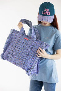 A person wearing a blue t-shirt and a teal cap with 'DOG LOVER' in pink text holds a large woven purple tote bag with fringe details against a plain white background.