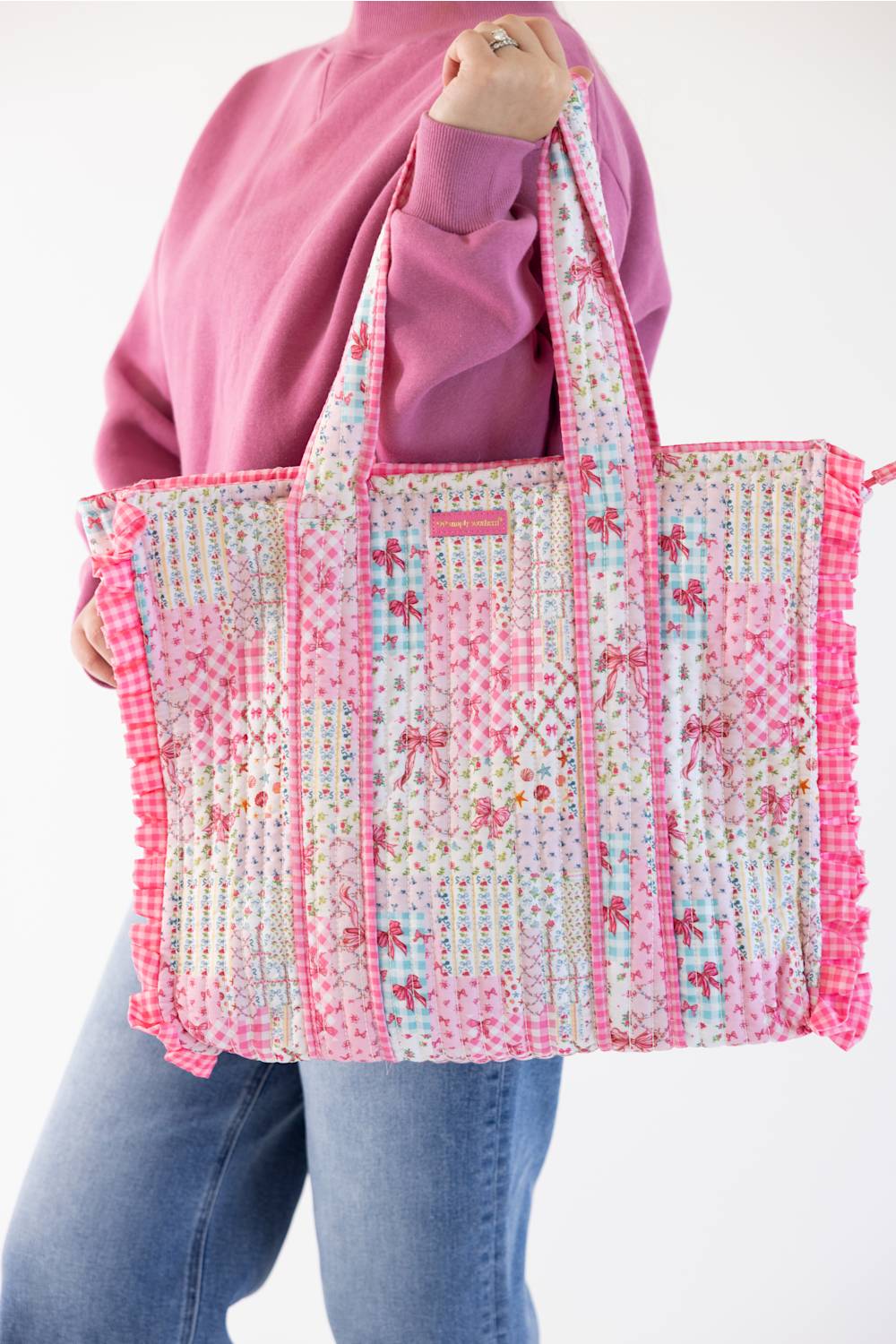 A person wearing a pink sweatshirt and blue jeans holds up a large quilted tote bag with pink gingham and floral patchwork panels, set against a plain white background.