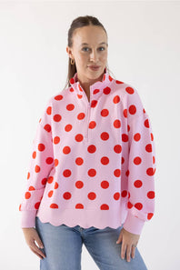 A woman wearing a pink long-sleeve quarter-zip pullover sweatshirt with a red polka dot pattern and a scalloped hem, paired with blue jeans, against a plain white background.