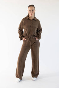 A woman modeling a matching brown two-piece lounge set consisting of a long-sleeve button-front top and wide-leg drawstring pants, photographed against a plain white studio background.