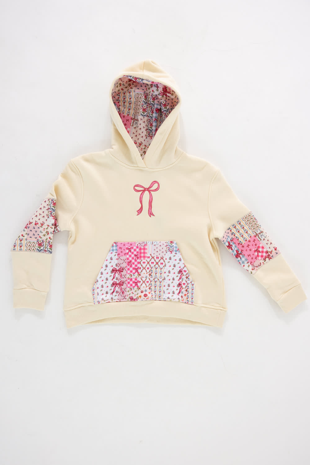 Front view of a girls' cream-colored pullover hoodie featuring an embroidered pink ribbon bow on the chest, a pink quilted patterned kangaroo pocket, patchwork floral elbow patches, and a patterned hood lining against a plain white background.