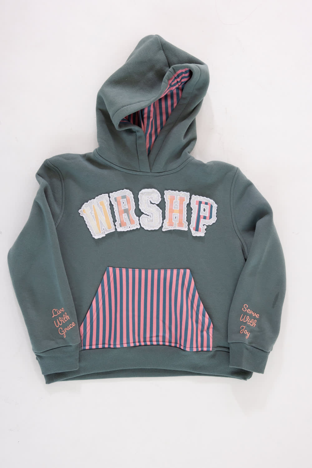Front view of a girls' gray pullover hoodie featuring patterned applique lettering that spells “WRSHP” across the chest, a pink and navy striped kangaroo pocket, a striped lined hood, and embroidered text on the sleeve cuffs reading “Live With Grace” and “Serve With Joy” against a plain light background.