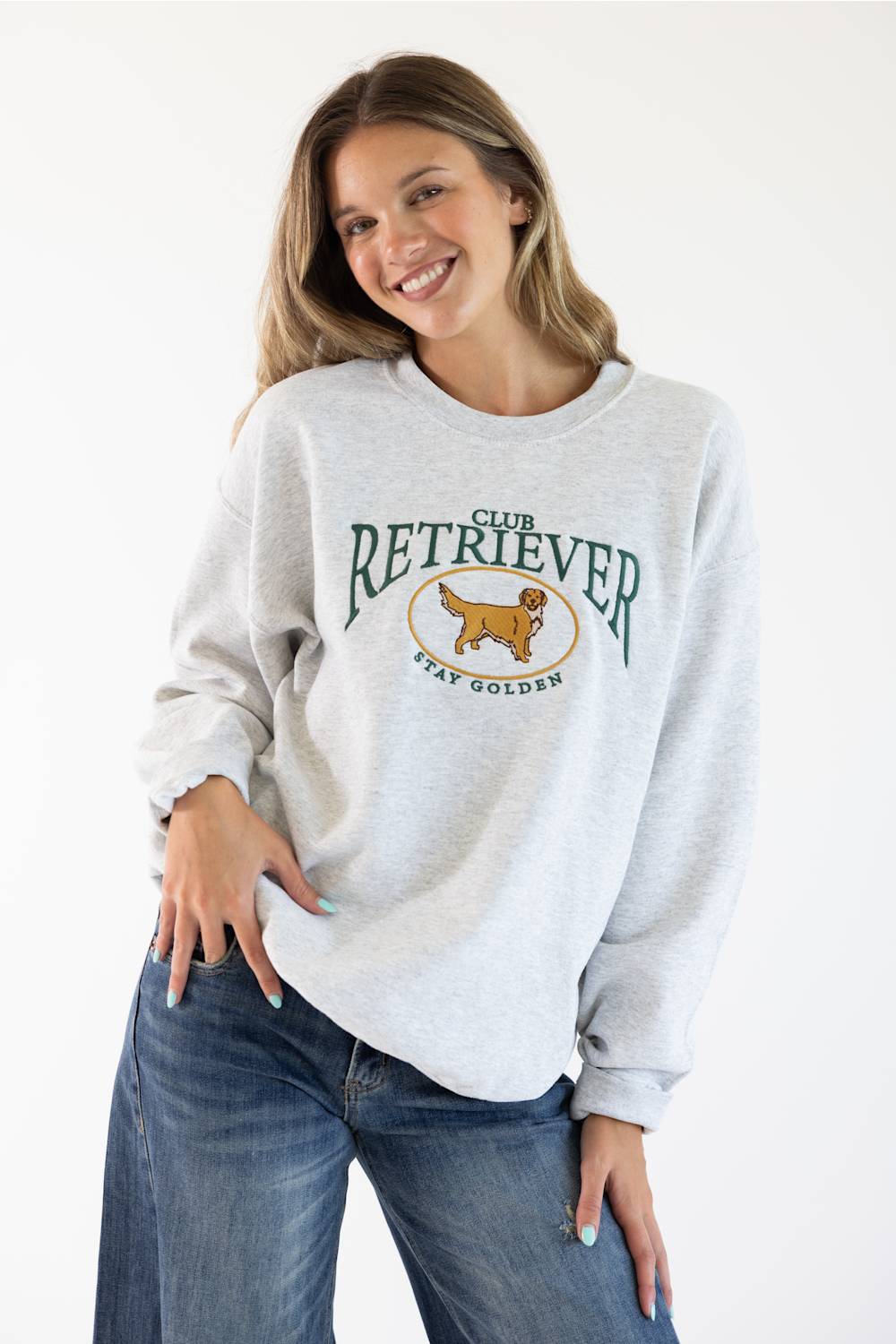 Front ¾ view of a smiling female model wearing the Sweet Claire Embroidered Club Retriever Sweatshirt in Ash heather grey, featuring green "Club Retriever Stay Golden" chest embroidery and relaxed fit paired with denim jeans.