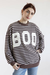 A woman posing in a brown and white horizontally striped sweatshirt with a large white BOO graphic on the front, paired with light blue jeans, against a plain white background.