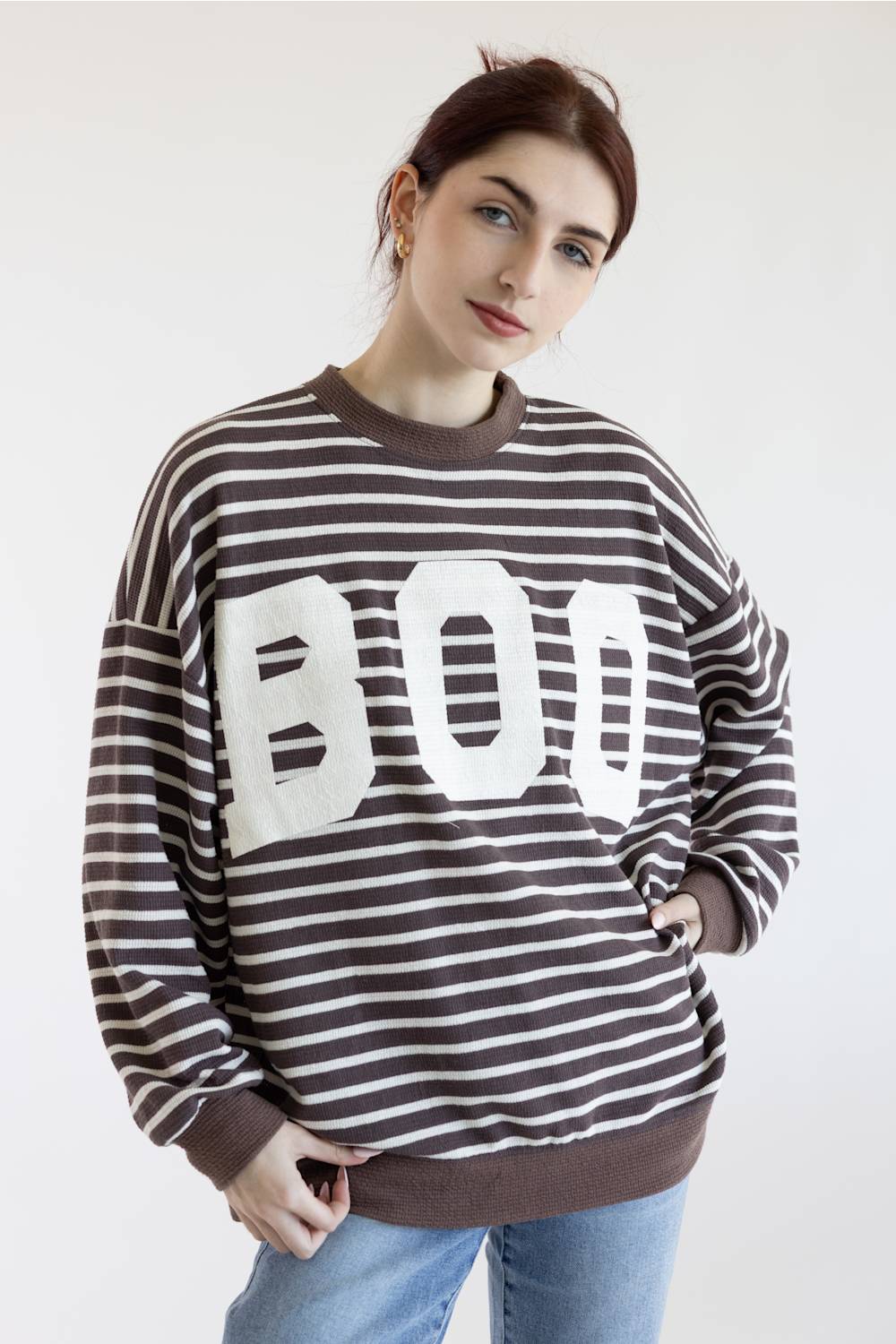 A woman posing in a brown and white horizontally striped sweatshirt with a large white BOO graphic on the front, paired with light blue jeans, against a plain white background.