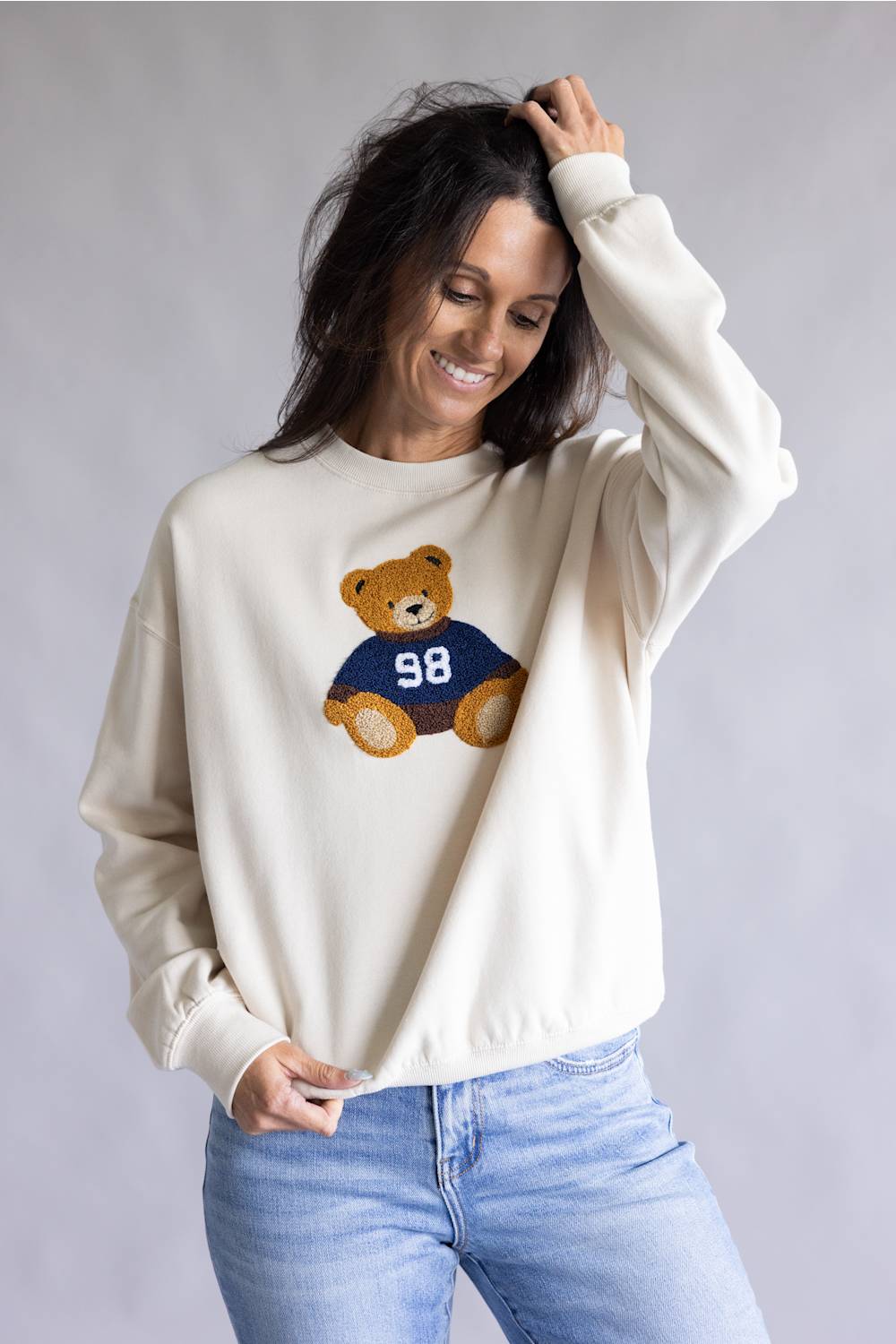 A woman wearing a cream-colored crewneck sweatshirt with a teddy bear graphic wearing a navy blue jersey with the number 98, paired with light blue jeans, against a neutral gray studio background.