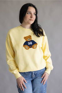 A woman wearing a pale yellow crewneck sweatshirt with a teddy bear graphic wearing a navy jersey with the number 98, paired with blue jeans, against a plain gray studio background.