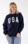 Front view of a blonde woman wearing a navy blue Reflex fleece crewneck sweatshirt with white "USA" block lettering across the chest and blue jeans.