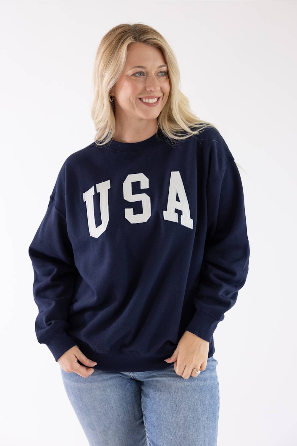 Front view of a blonde woman wearing a navy blue Reflex fleece crewneck sweatshirt with white "USA" block lettering across the chest and blue jeans.