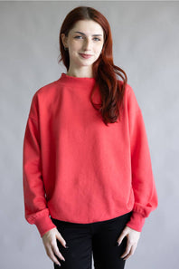 A woman with long red hair wearing a loose-fitting coral red sweatshirt and black pants, standing against a plain gray studio background.