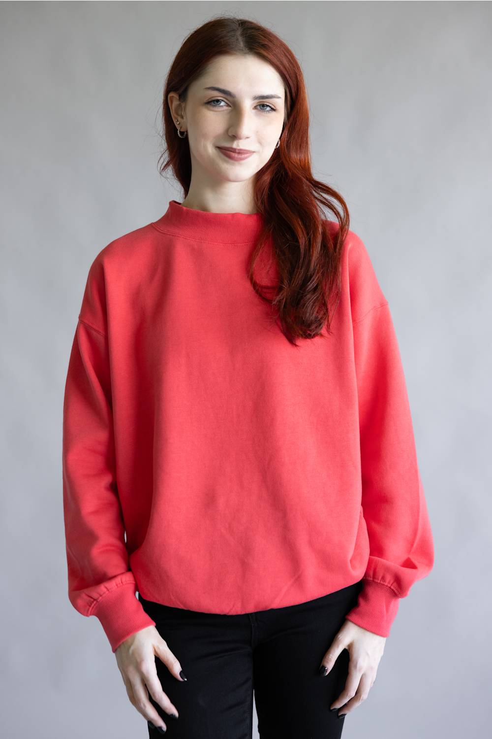 A woman with long red hair wearing a loose-fitting coral red sweatshirt and black pants, standing against a plain gray studio background.