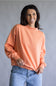 A woman wearing a peach-colored crewneck sweatshirt and light blue jeans, standing against a neutral gray studio background.