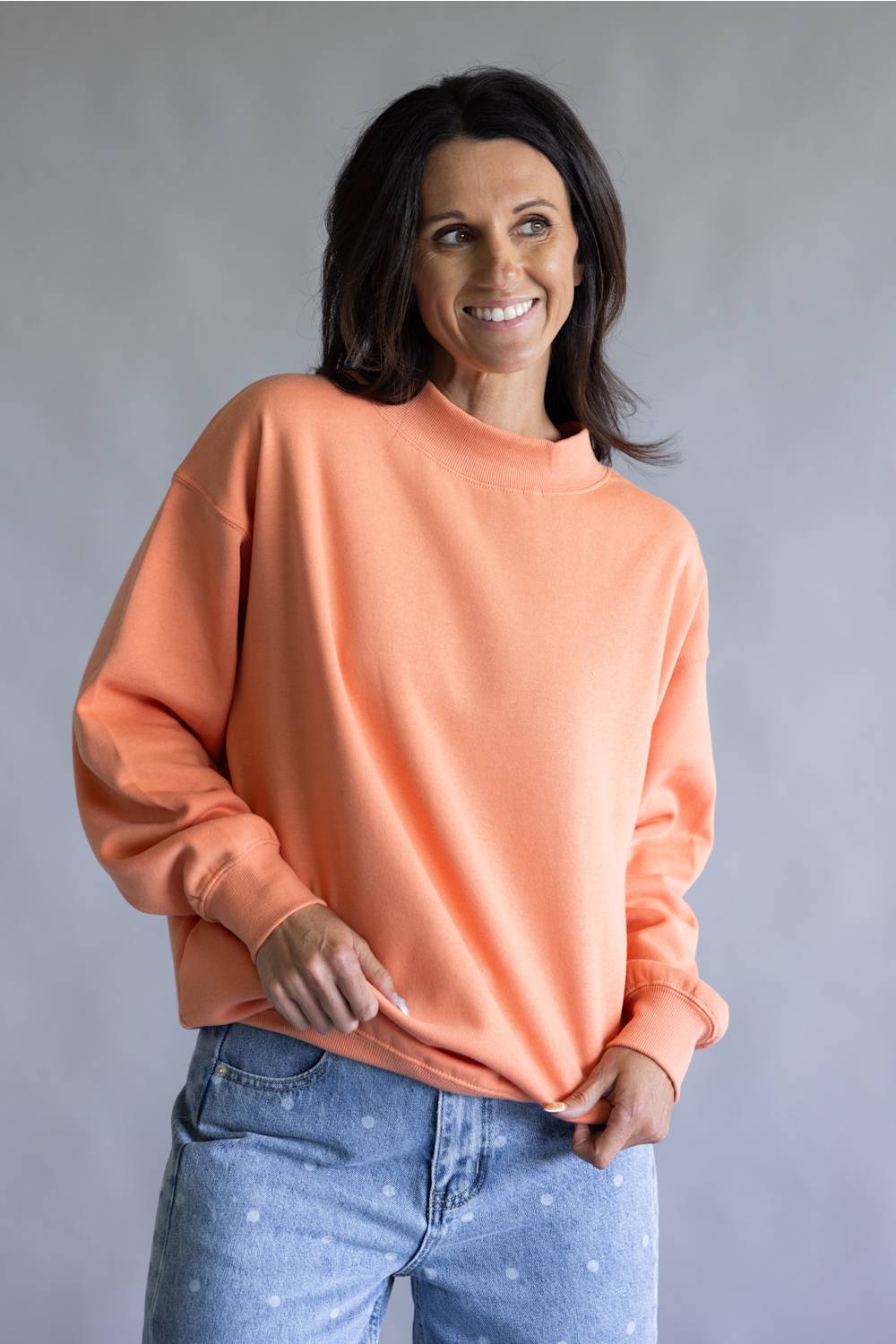 A woman wearing a peach-colored crewneck sweatshirt and light blue jeans, standing against a neutral gray studio background.