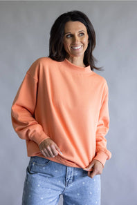 A woman wearing a peach-colored crewneck sweatshirt and light blue jeans, standing against a neutral gray studio background.