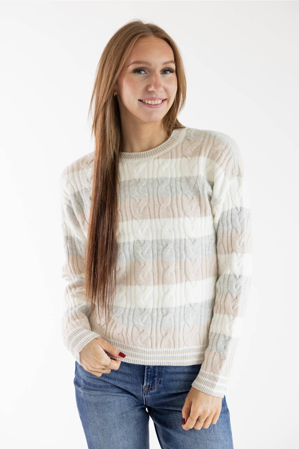 A woman with long light brown hair wearing a cream, beige, and light gray striped cable-knit sweater and blue jeans, standing against a plain white studio background.
