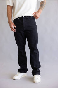A person wearing black straight-leg jeans, a white textured t-shirt, and white sneakers, standing against a light gray studio background.