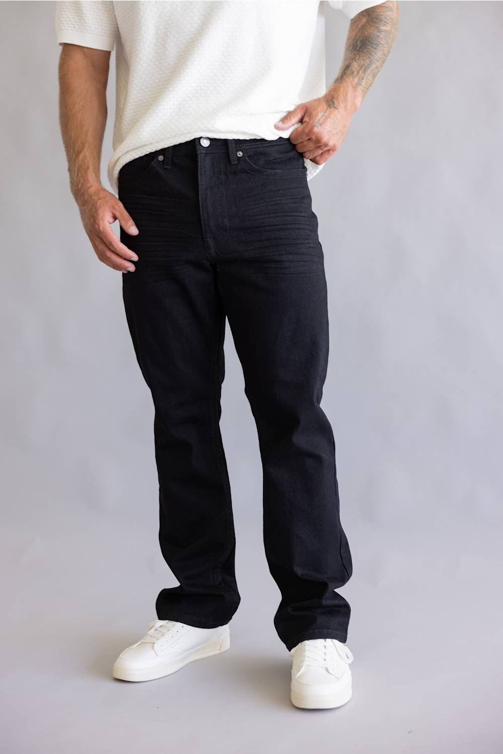A person wearing black straight-leg jeans, a white textured t-shirt, and white sneakers, standing against a light gray studio background.