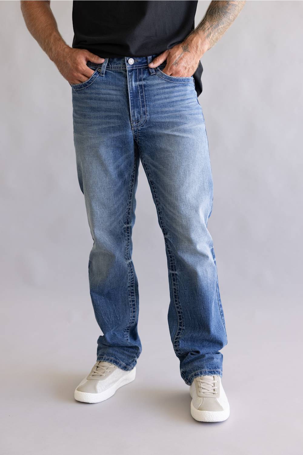 A person wearing light-to-medium wash blue jeans with a black t-shirt and white sneakers, standing against a light gray studio background.
