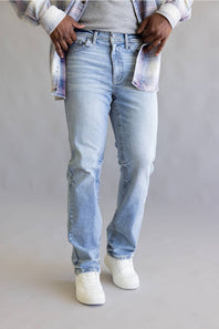 A person wearing light-wash blue jeans, a gray top, and white sneakers, standing against a plain light gray studio background.