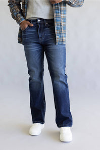 A person wearing dark blue straight-leg denim jeans, styled with a blue and tan plaid flannel shirt over a light gray t-shirt and white sneakers, against a light gray studio background.