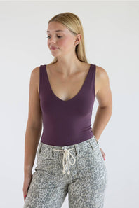 A woman wearing a sleeveless deep purple scoop-neck tank top and gray leopard-print pants with a white drawstring, standing against a plain white studio background.