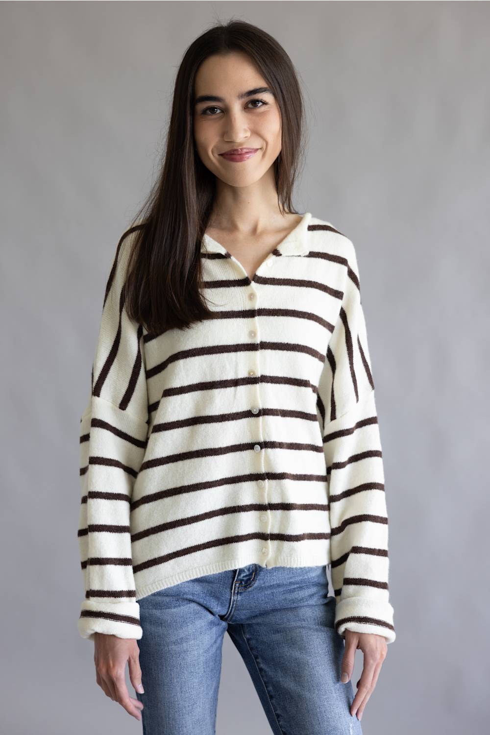 A woman wearing a cream-colored knit cardigan with dark brown horizontal stripes, paired with blue jeans, standing against a neutral gray studio background.
