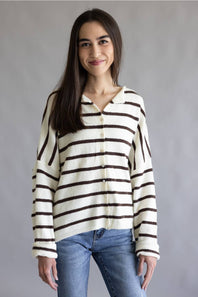 A woman wearing a cream-colored knit cardigan with dark brown horizontal stripes, paired with blue jeans, standing against a neutral gray studio background.