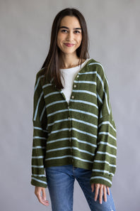 A woman modeling an olive green and light blue striped knit cardigan over a white top and blue jeans, standing against a neutral gray studio background.