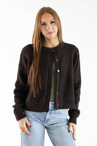 A woman modeling a dark brown knit cardigan over an olive green top and light blue jeans, photographed against a plain white studio background.