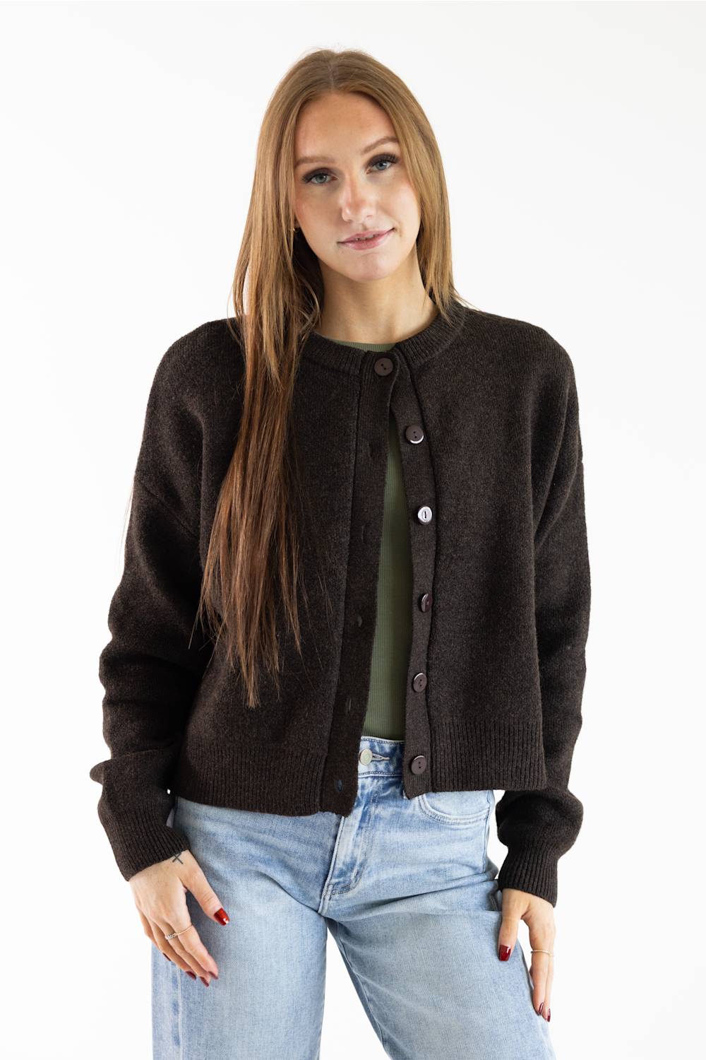 A woman modeling a dark brown knit cardigan over an olive green top and light blue jeans, photographed against a plain white studio background.