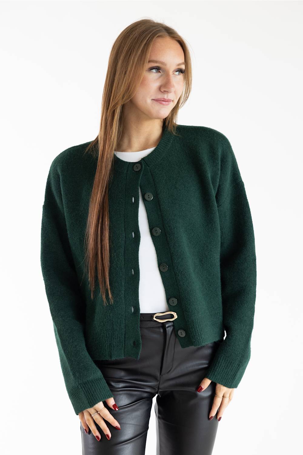 A woman modeling a dark forest green knit cardigan with dark buttons, worn open over a white t-shirt and black faux-leather pants, against a plain white studio background.
