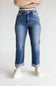 A person wearing medium-wash blue jeans with a striped waistband, cuffed hems, and light beige slip-on shoes, shown from the waist down against a plain light gray studio background.