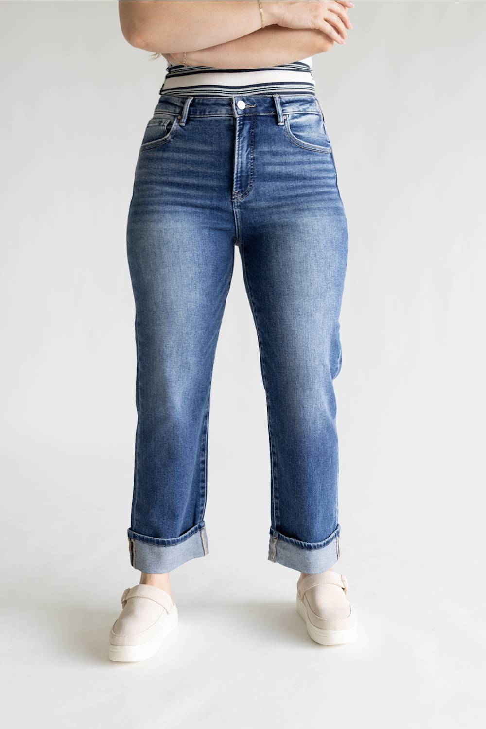 A person wearing medium-wash blue jeans with a striped waistband, cuffed hems, and light beige slip-on shoes, shown from the waist down against a plain light gray studio background.