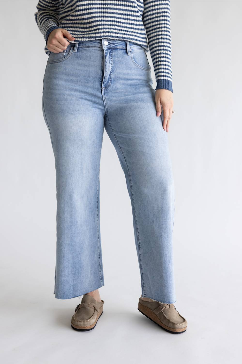 A person wearing light-wash blue straight-leg jeans, a navy and white striped long-sleeve top, and brown suede loafers, standing against a plain light gray studio background.